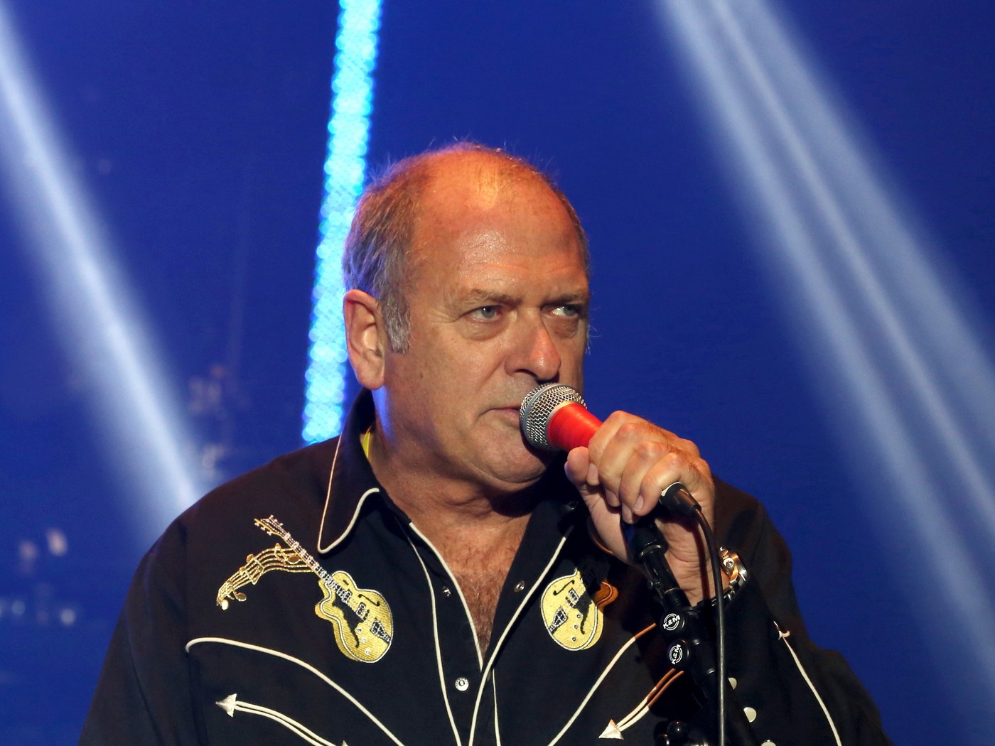 Spike Edney performing live on stage, singing into a handheld microphone under blue stage lighting